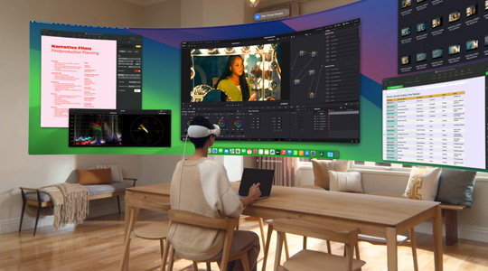 Eine Person, die die Apple Vision Pro trägt, sitzt an einem Schreibtisch mit einem geöffneten 13" MacBook Pro, und ein gebogenes, ultrabreites Display mit den Inhalten ihres Bildschirms wird in den Raum projiziert