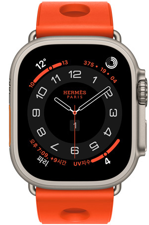 Apple Watch Hermès Ultra 3 Circulaire 아날로그 시계 페이스에 (시, 분, 초침, 컴플리케이션: 날씨, 활동 링, 파리 시간 및 자외선 지수), 내추럴 색상 티타늄 케이스, 오른쪽 옆면 외관, Digital Crown, 돌출된 측면 버튼, 오랑쥬 네옹(밝은 오렌지) 색상의 Scub’H Diving 밴드(중앙에 구멍이 뚫린 성형 고무 소재)와 연결되어 있다.
