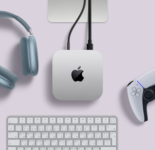 لقطة علوية لجهاز Mac&nbsp;mini فضي اللون، مع سلكين متصلين بالمنافذ الخلفية، وبجواره سماعات AirPods&nbsp;Max باللون الأزرق، ولوحة مفاتيح ماجيك باللون الفضي، ووحدة تحكم ألعاب لاسلكية