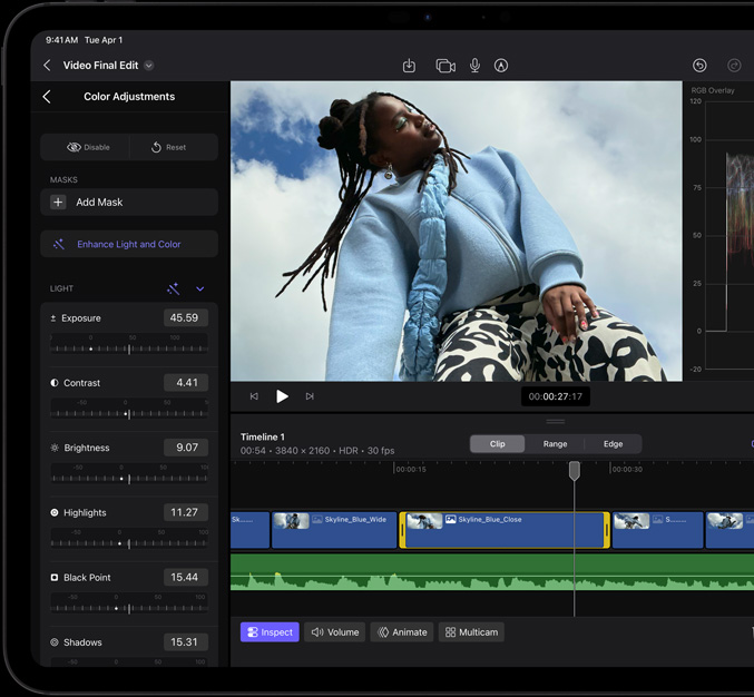 iPad Pro, front exterior, black display bezel, display showing video editing app with a picture of a model in a blue coat and black and white printed trousers, colour adjustment panel open on the left with various video effects and sliders, timeline at the bottom with multiple video clips and audio tracks