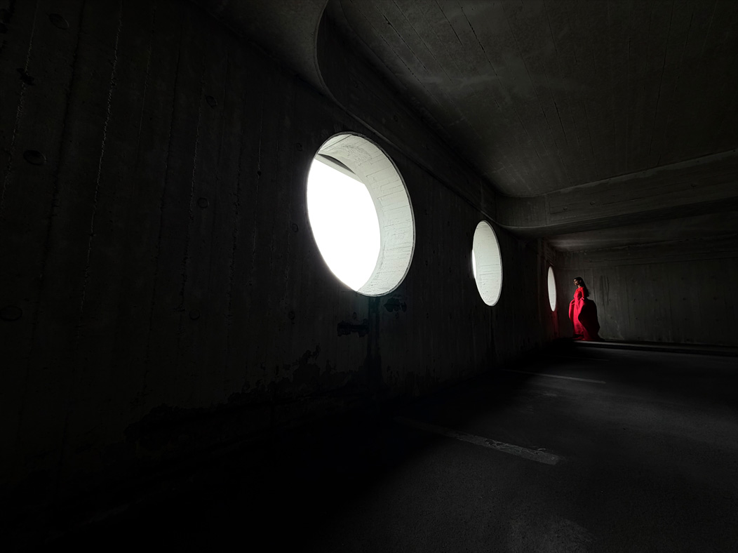 Eine Frau in einem langen roten Kleid steht in einem leeren Raum und wird fotografiert. Die Fotos App wechselt durch die Brennweiten, um die Möglichkeiten des Pro Kamera-Systems zu zeigen, hier: 13 mm